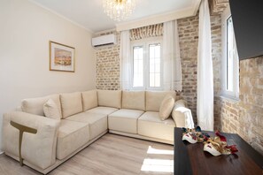 A room with a beige sectional sofa, a wooden table with decorative items, a chandelier, a framed picture on the wall, and a brick wall. - Borgo Veneziano by Corfuescapes (Kerkira)