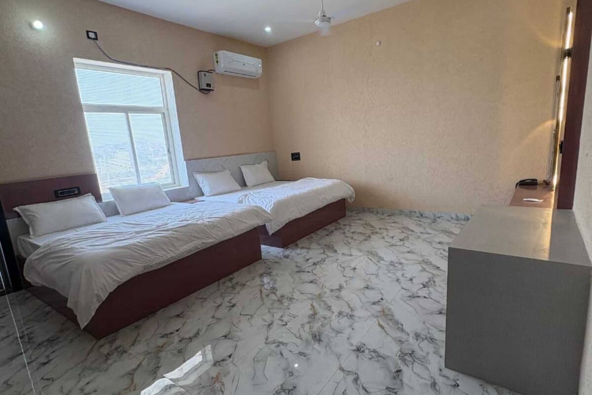 Habitación de hotel con dos camas, ventilador de techo, equipo de aire acondicionado, ventana con persianas y piso de mármol.