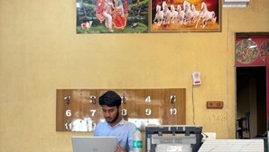 Una reception con una persona che lavora al computer portatile, un telefono a muro e una porta con un cartello.