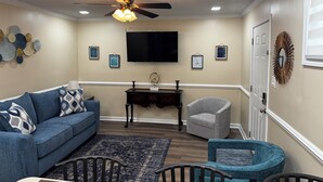 Una sala de estar con ventilador de techo, televisor de pantalla plana, sofá, sillón, mesita pequeña y un adorno en la pared.