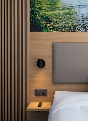 Une chambre d’hôtel moderne avec une tête de lit en bois, une lampe fixée au mur, une prise de courant et une télécommande posée sur une table de chevet en bois.