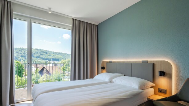 Une chambre d’hôtel avec un grand lit, une chaise, une table de chevet et une vue sur un paysage verdoyant, visible à travers une porte-fenêtre coulissante.