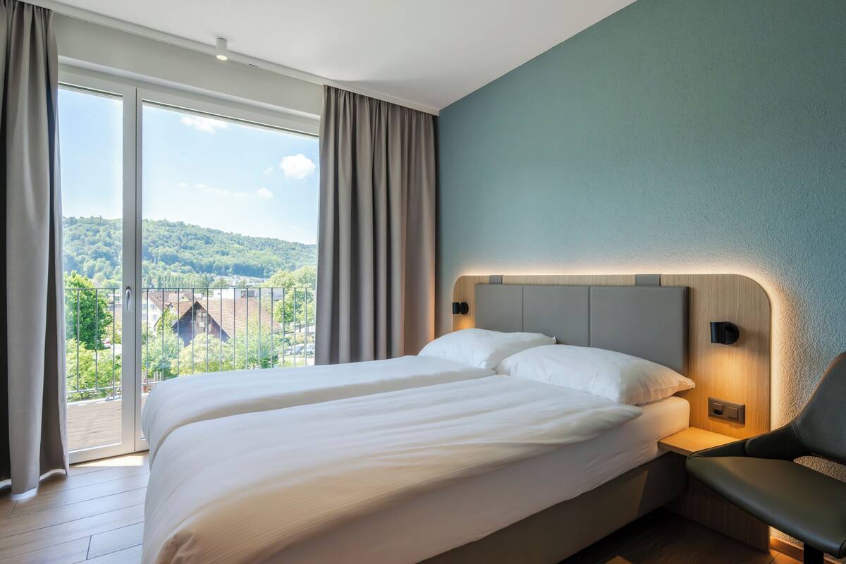 Hotelkamer met een groot bed, een stoel, een nachtkastje en uitzicht op een groen landschap door een schuifdeur.