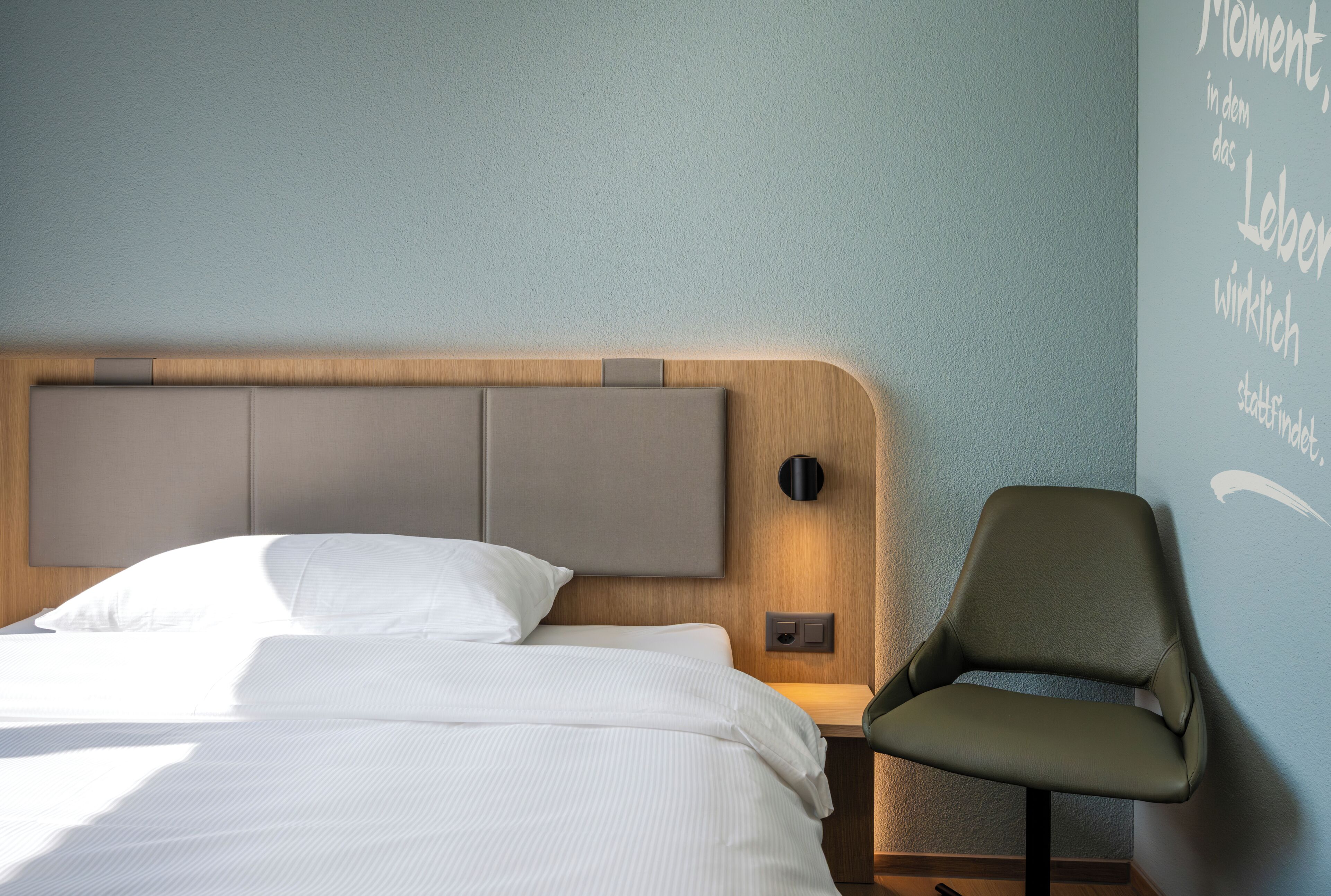 Ein Hotelzimmer mit einem Bett, einem Sessel, einer Wandleuchte und einer Wand mit Schrift.