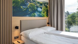 Une chambre d’hôtel moderne dotée d’un grand lit, d’une table de chevet, d’une lampe fixée au mur et d’une fresque murale représentant un paysage urbain et une rivière.