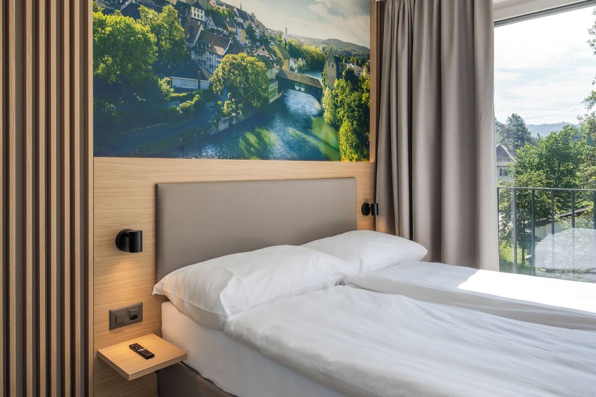 Een moderne hotelkamer met een groot bed, een nachtkastje, een wandlamp en een sfeervol muurdecoratie van een stad en rivier.