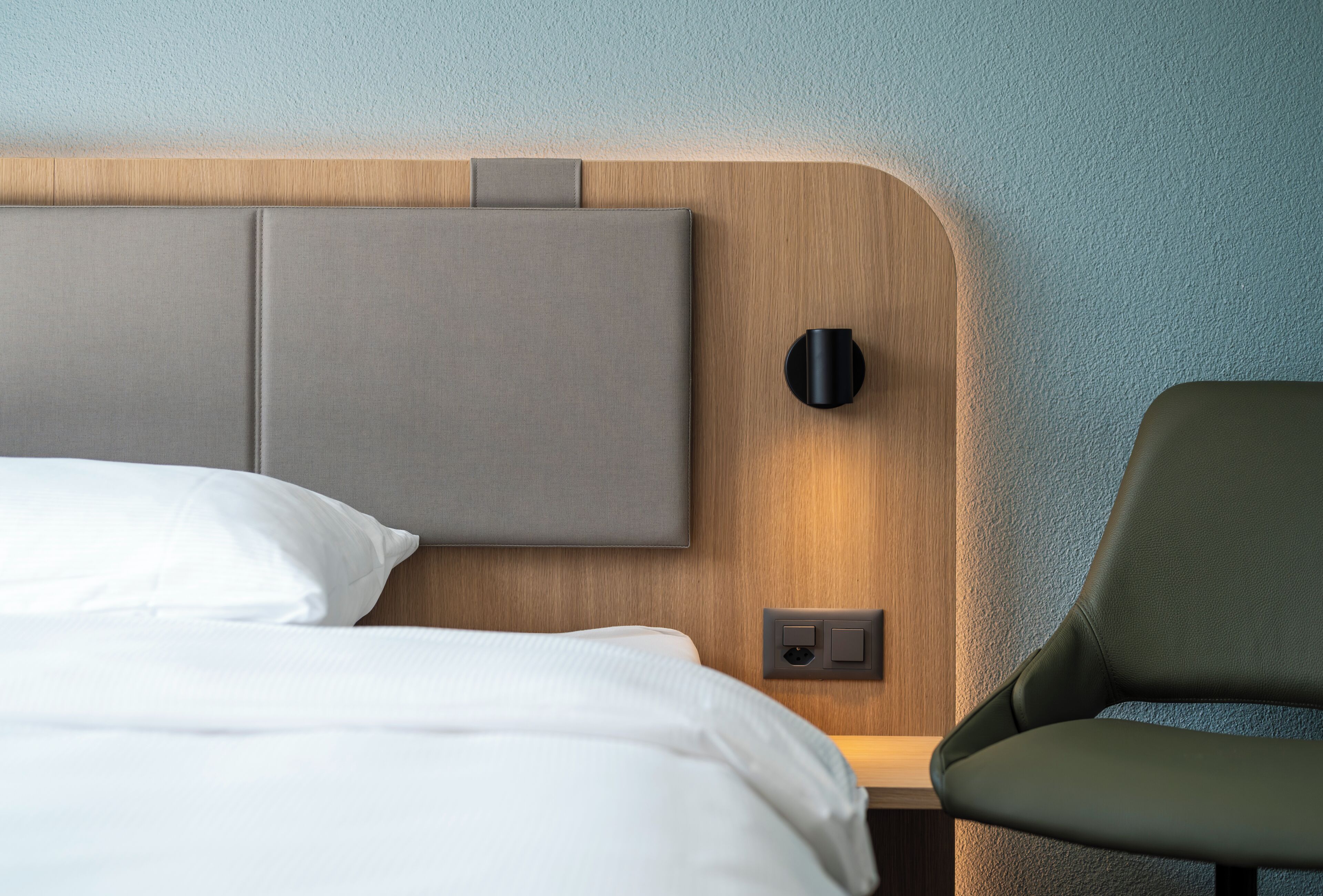 Un lit bien fait, avec du linge de lit blanc, une tête de lit en bois, une lampe fixée au mur et une chaise verte.
