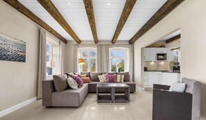 A modern living room with a sectional sofa, a small table, and a kitchen in the background. - Holiday apartment Lille Hus 1 in St. Peter-Ording: 95 m² with terrace (Sankt Peter-Ording)