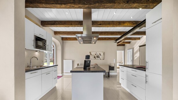 Une cuisine moderne avec des meubles blancs, un îlot central, une hotte de cuisine en acier inoxydable et des poutres apparentes en bois.