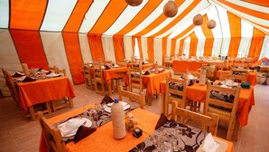 Un área de comedor con carpas, mesas dispuestas para una comida, con manteles anaranjados y sillas de madera.