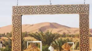 Paisaje desértico con un gran marco de madera ornamentado, una estructura blanca y una lámpara tradicional.