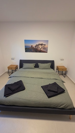 Un lit recouvert d’une couverture verte, avec deux oreillers et deux couvertures pliées. Deux tables de chevet, dotées de côtés en bois et de pieds en métal noir, se trouvent de chaque côté du lit. Un tableau encadré est accroché au-dessus du lit.