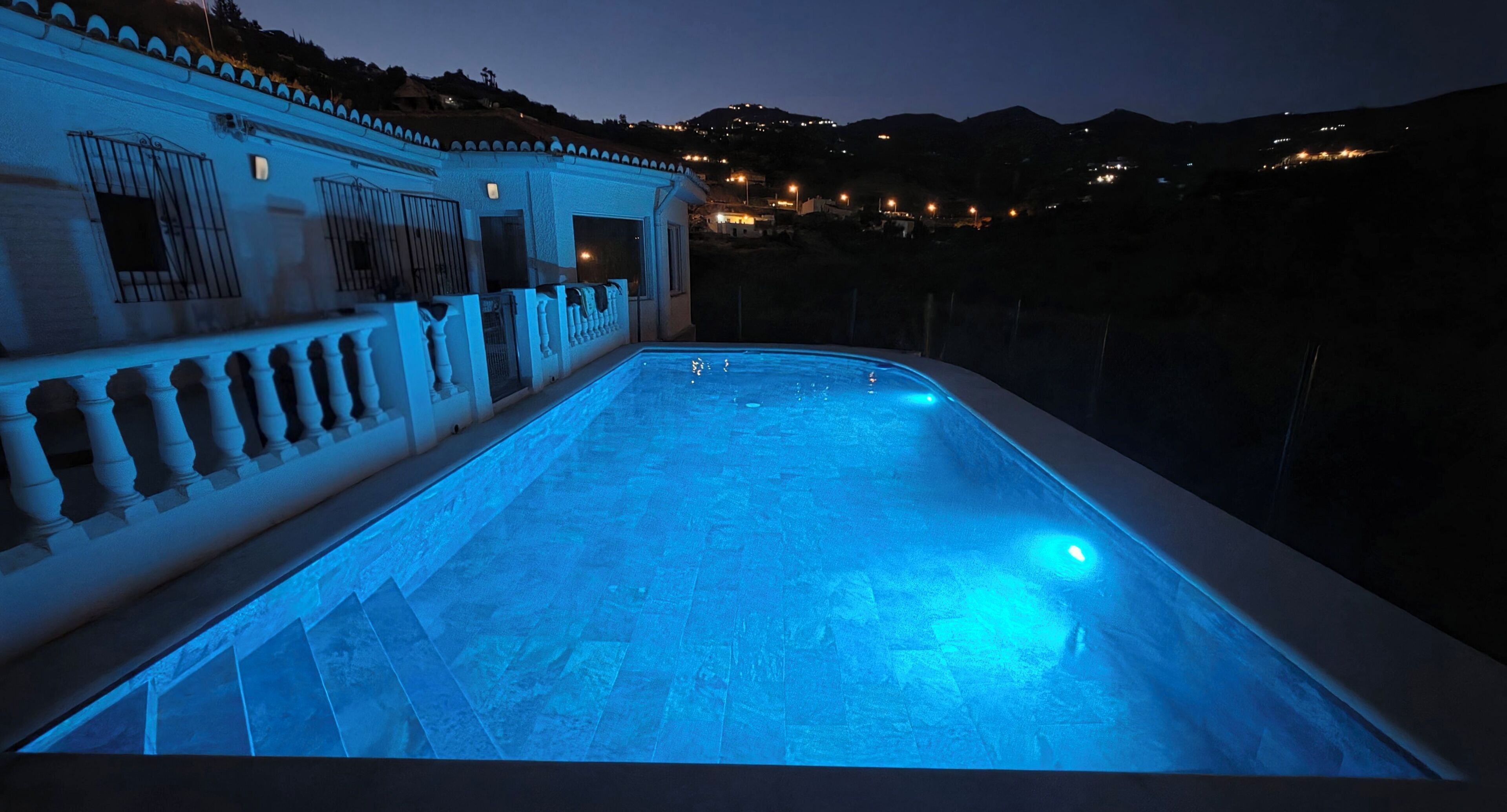 A swimming pool with a blue-lit edge, surrounded by a white wall with decorative railings and a view of a hillside with distant lights.