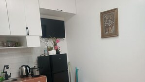 Uma cozinha compacta com armários brancos, uma geladeira preta, uma pia e uma cama com colcha bege.