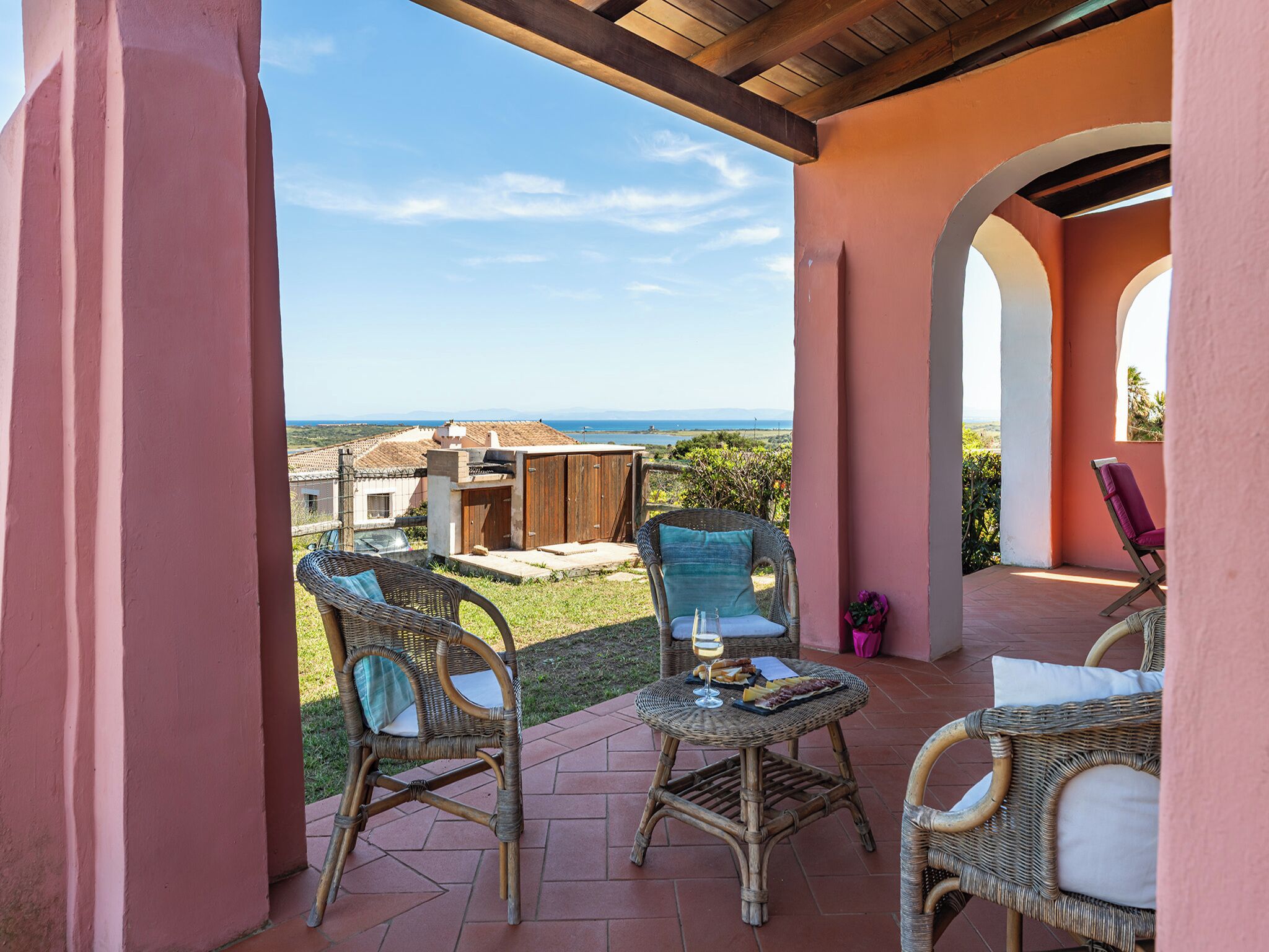 A covered patio with wicker furniture, a table with food and drink, and a view of the sea and distant buildings.