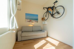 Quarto com sofá, bicicleta fixada na parede, pintura de uma cena de praia e janela com cortinas.
