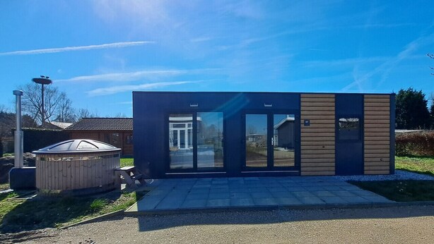 A modern building with a glass entrance, a wooden structure to the left, and a clear sky. - Cube 97 | 4 persoons chalet met hottub (Zennewijnen)