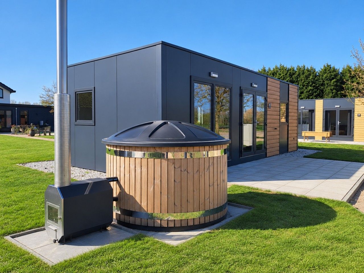 A modern building with a wooden hot tub and a metal heater in the foreground, and a patio area with a bench.