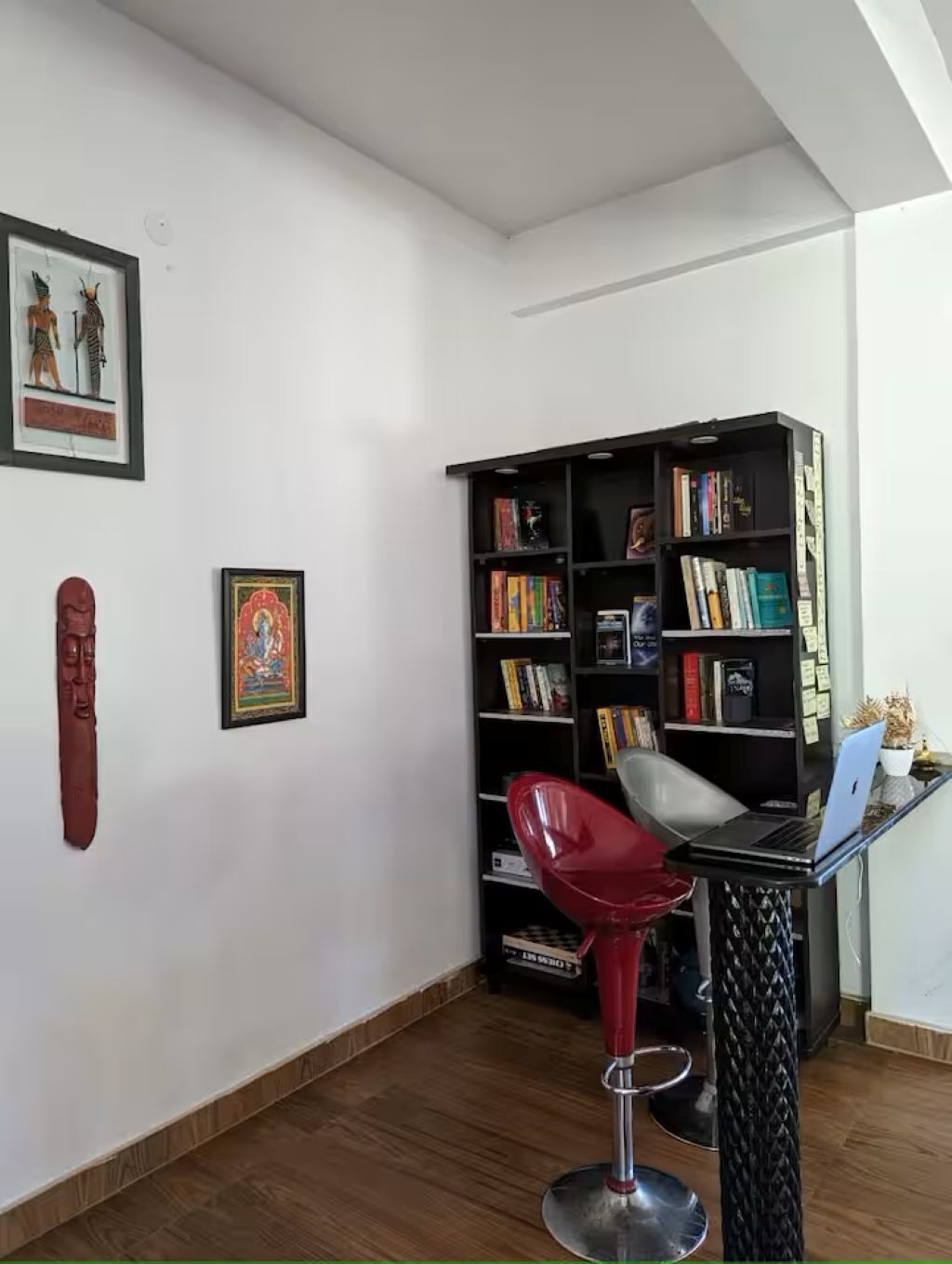 A room with a wooden floor, a red chair, a black bookshelf, and two framed pictures on the wall.