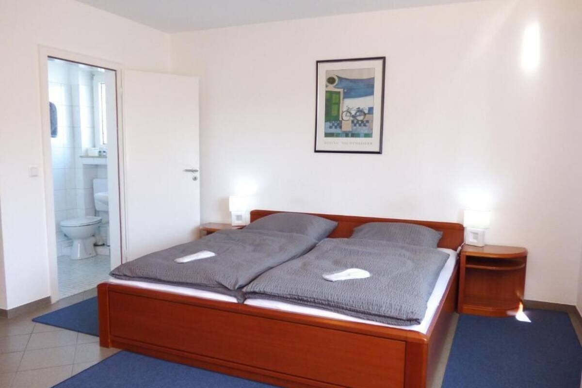A bedroom with a wooden bed, two bedside tables, a framed picture on the wall, and a bathroom visible through an open door.