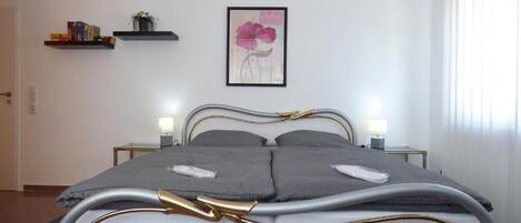 A modern bedroom with a bed, bedside tables, a wall-mounted shelf with books, a framed floral artwork, and a window with blinds.