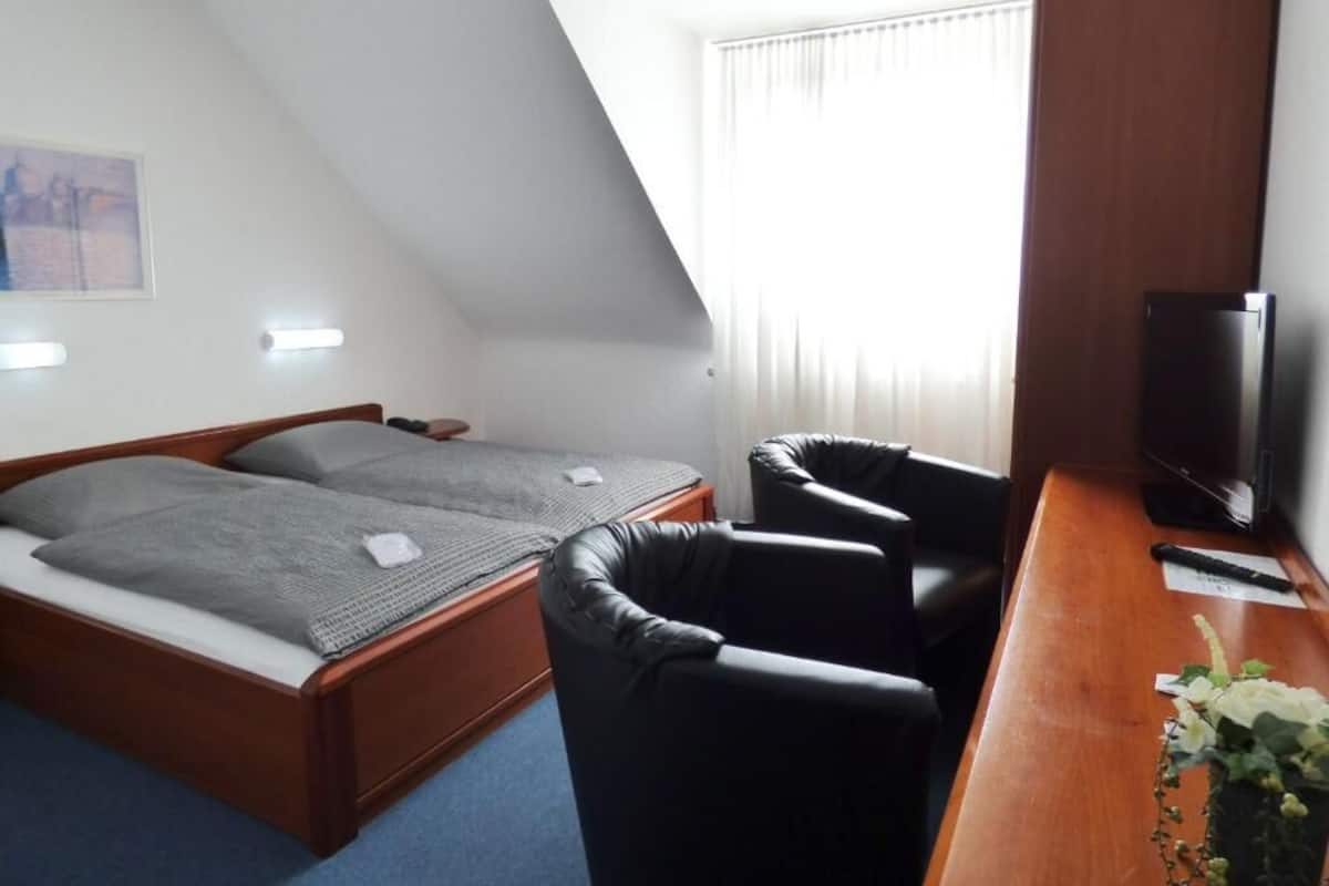A bedroom with a bed, a desk with a chair, a television, and a window with blinds.