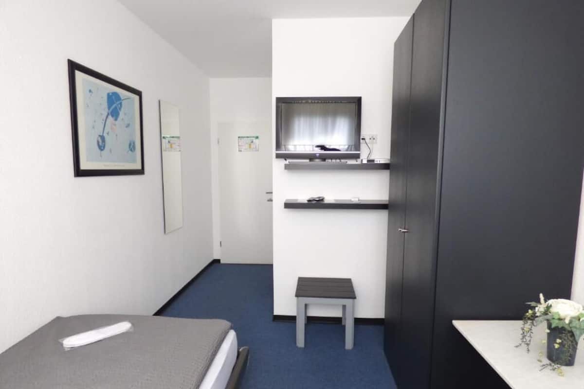 A small, modern room with a bed, a desk, a chair, a small table, a cabinet, and a framed picture on the wall.