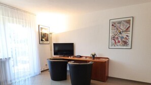 Una stanza con una TV a schermo piatto, una scrivania in legno, due sgabelli neri, un tappetino da yoga blu e un grande quadro astratto appeso al muro.