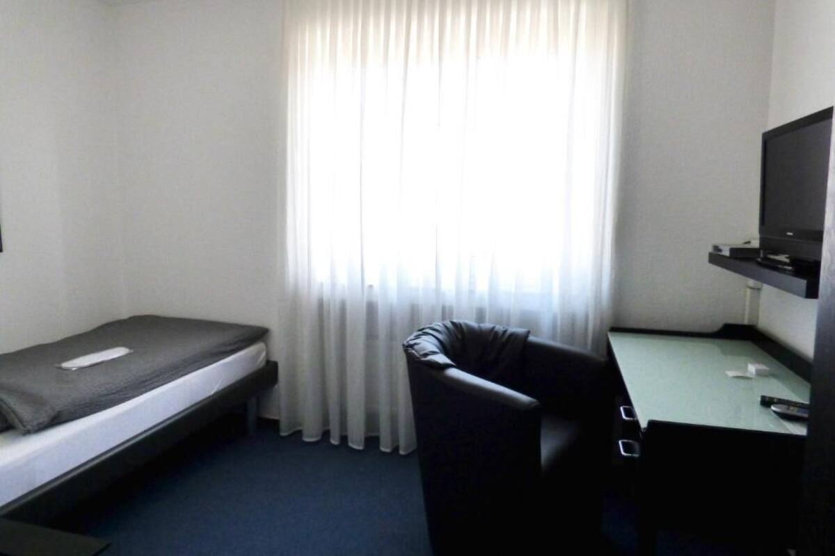 A small hotel room with a single bed, a desk with a chair, a television, and a window with curtains.