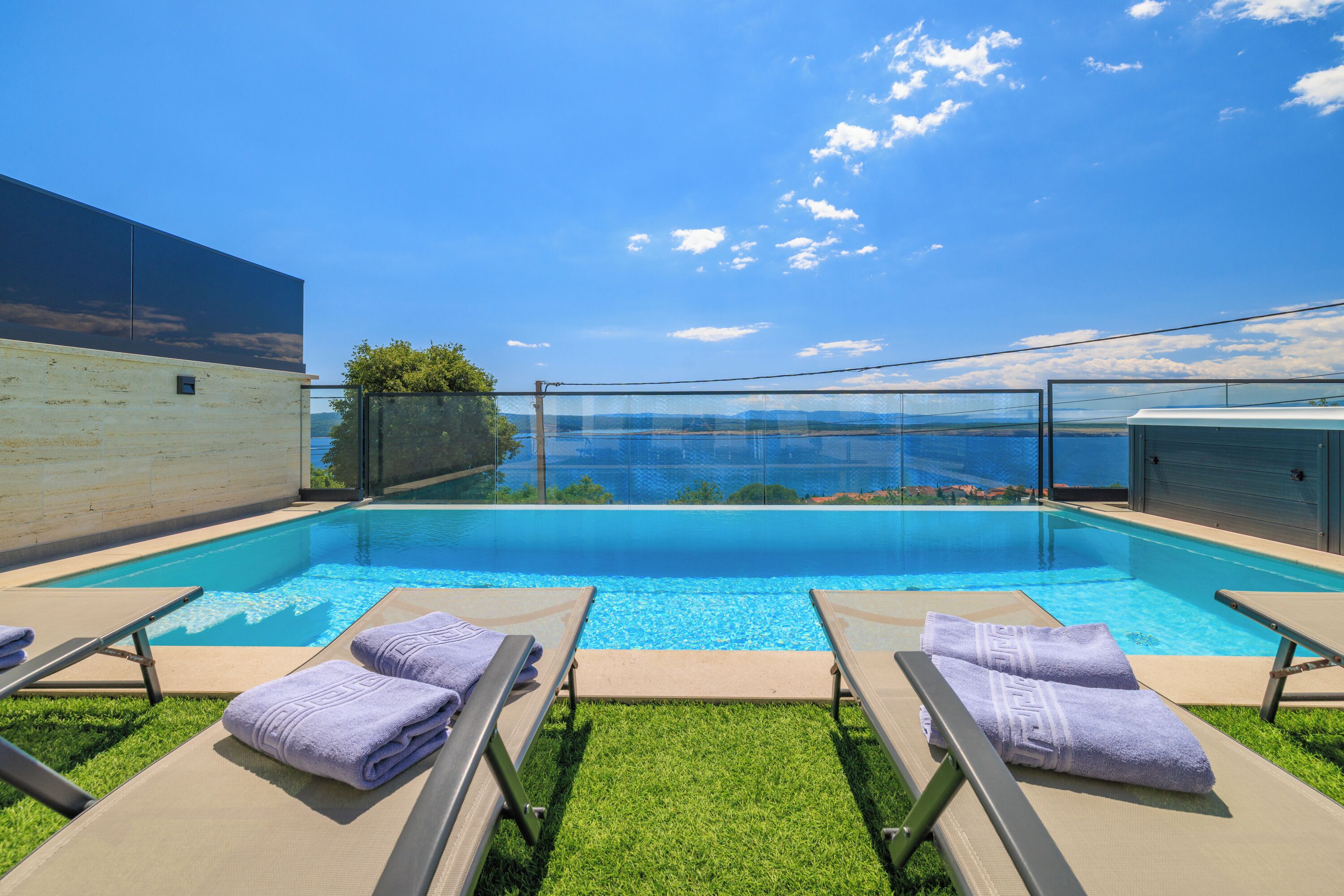 A swimming pool with lounge chairs and towels, surrounded by a clear sky and a view of the sea.