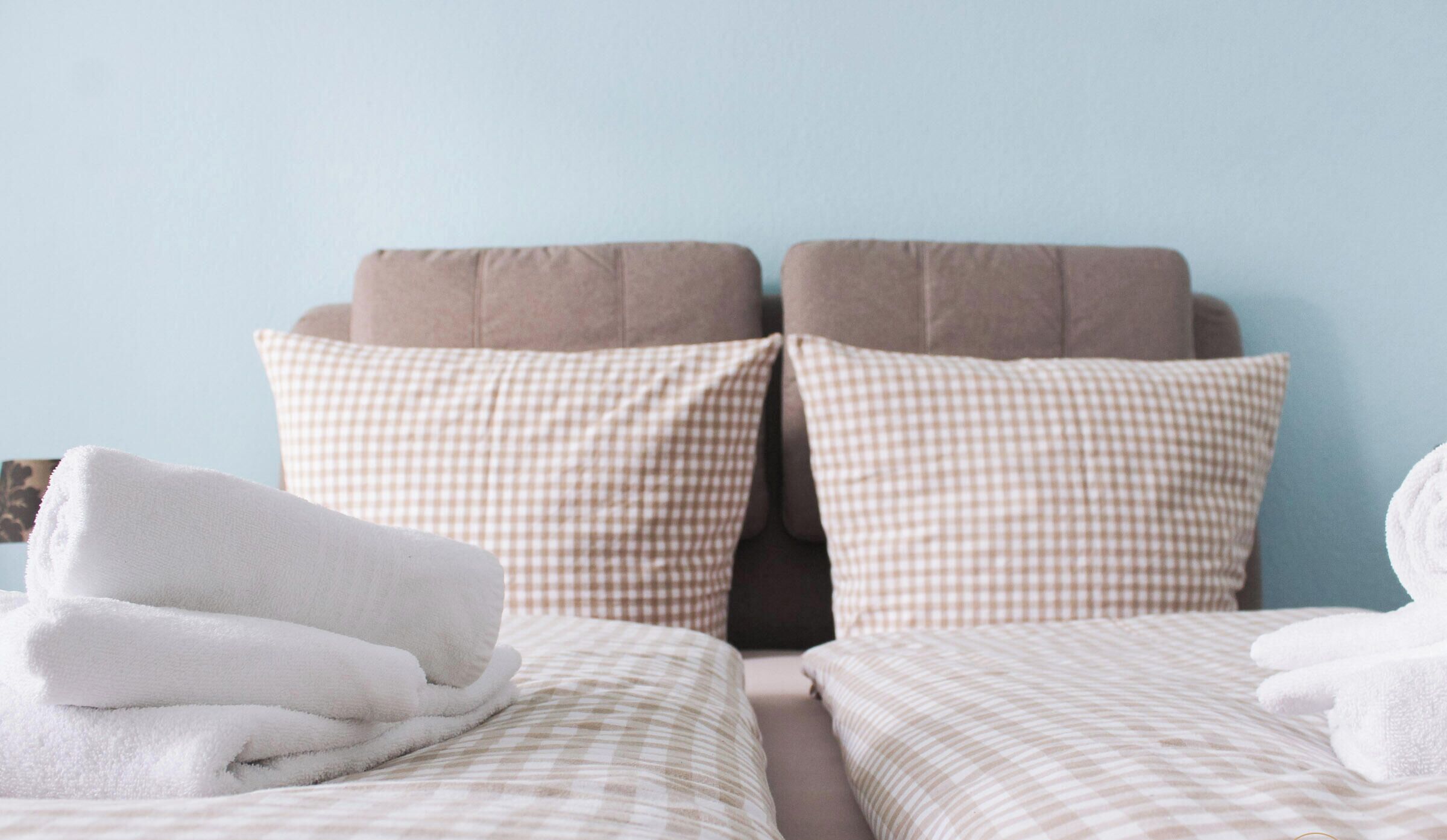 A bed with checkered bedding and two pillows, a folded white towel, and a light blue wall.