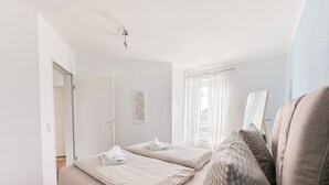 A bedroom with two beds, a window with curtains, a door, and a wall-mounted light fixture. - Vacation apartment Marina Liebe directly in the City Marina Cuxhaven (Cuxhaven)