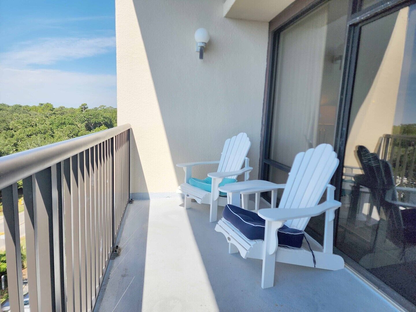 A balcony with two white Adirondack chairs, a small table, and a view of greenery.
