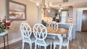 Un comedor con una mesa de madera y sillas blancas, un candelabro y una cocina al fondo.