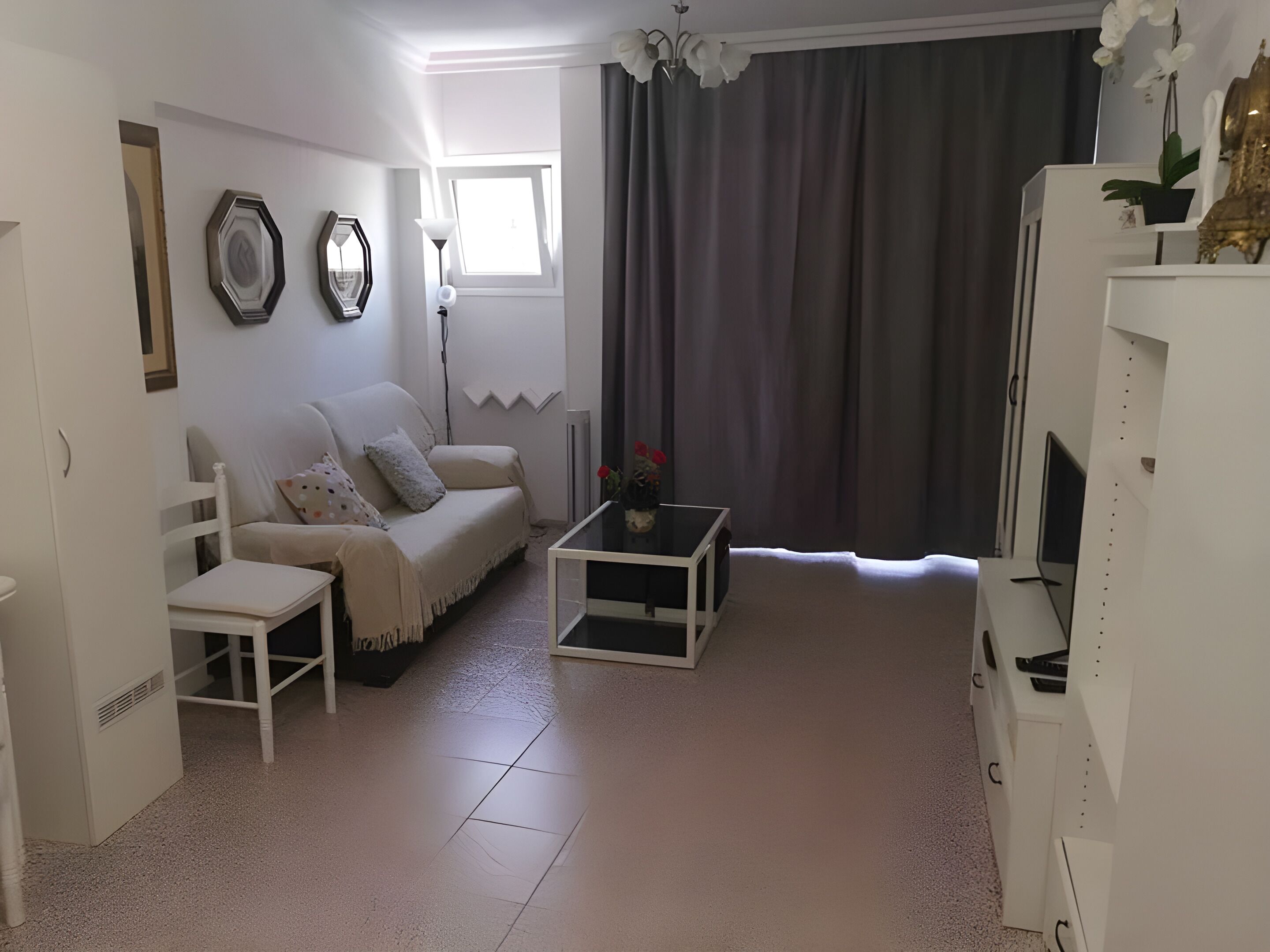 Una sala de estar con un sofá blanco, una mesita con un jarrón, un armario blanco y un ventanal con cortinas.