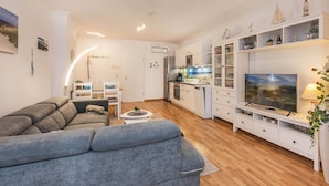 Ett modernt vardagsrum med en grå soffa, ett matbord och en köksdel med vita skåp och en inbyggd ugn.