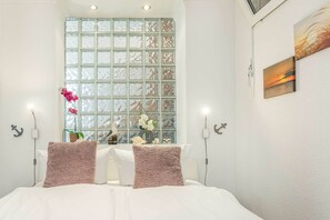 Ein Hotelzimmer mit großem Fenster, einem Bett mit zwei Kissen, einer Blumenvase und einem Gemälde an der Wand.