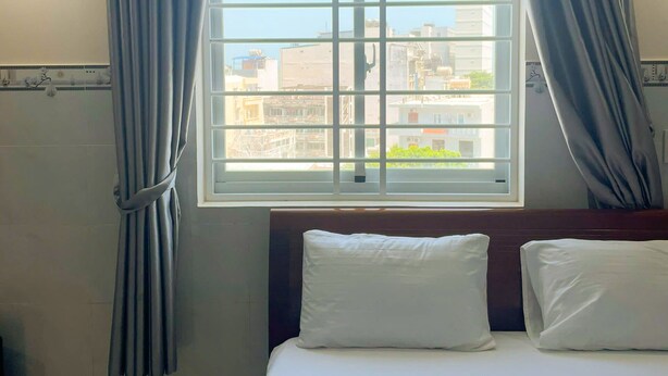 A hotel room with a large window, curtains, a bed with pillows, and a view of buildings outside. - Sunny Hotel Vung Tau (Ho Chi Minh City)