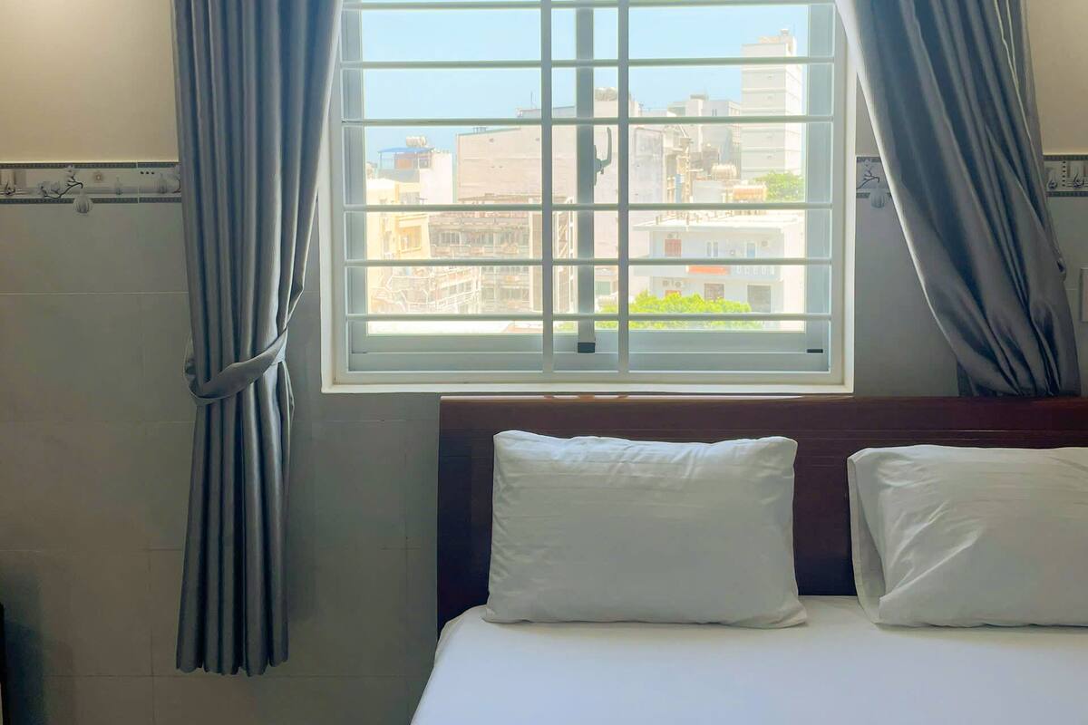 A hotel room with a large window, curtains, a bed with pillows, and a view of buildings outside.