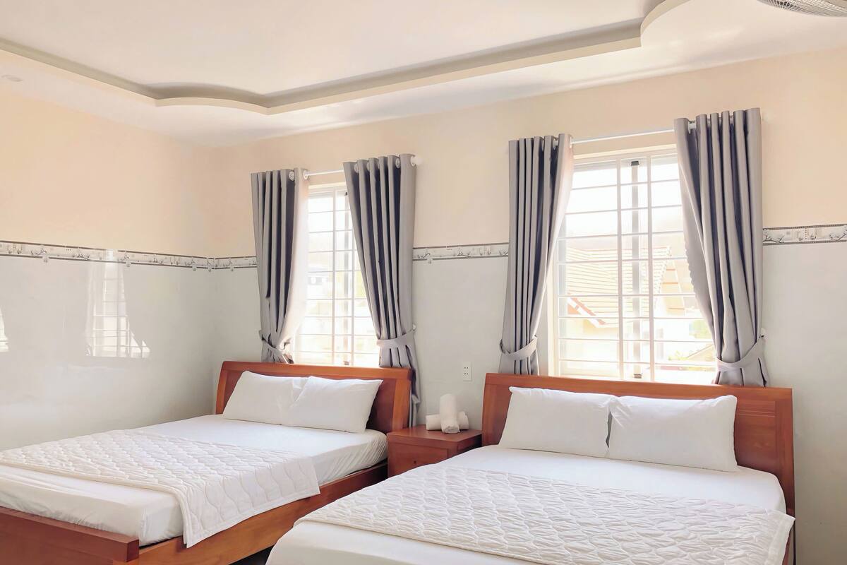 A hotel room with two beds, a window with curtains, a ceiling fan, and a nightstand with a lamp.