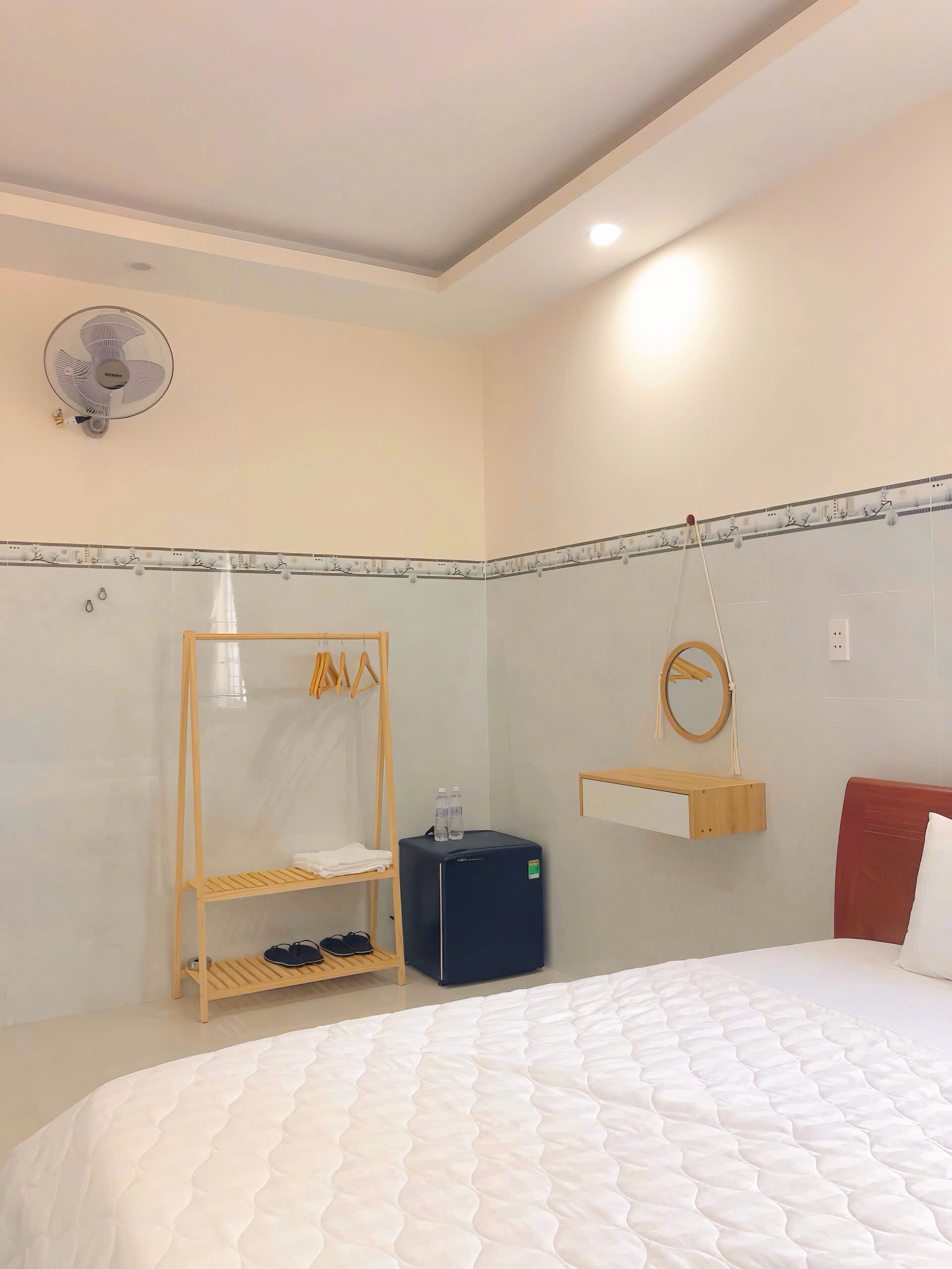 Une chambre avec un lit, une étagère en bois, une petite glacière noire, un ventilateur suspendu et un miroir fixé au mur.