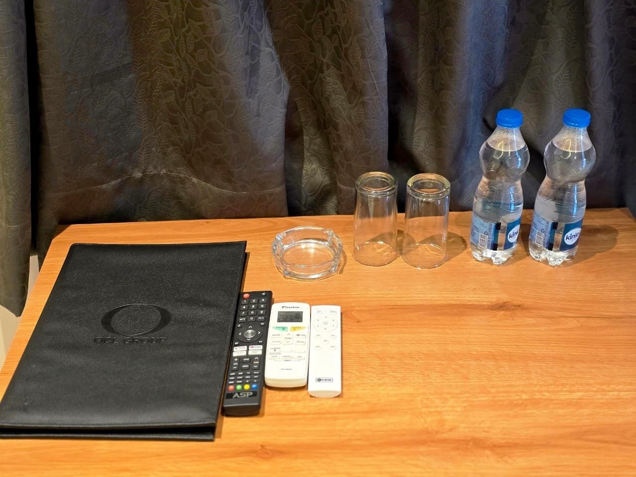 A wooden table with a black laptop, two remotes, two glass tumblers, and two bottles of water.