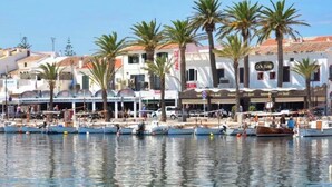 A waterfront area with boats docked, palm trees, and buildings with signage. - Casa Major by 3 Villas Menorca (Fornells)