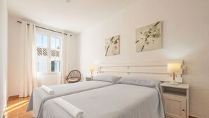 A bedroom with two beds, a nightstand with a lamp, a chair, and artwork on the wall. - Casa Major by 3 Villas Menorca (Fornells)