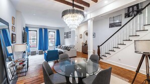 Una sala de estar moderna con una mesa de comedor de vidrio, sillas grises, una araña, una escalera y un sofá azul.