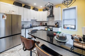 Une cuisine moderne avec des meubles blancs, un réfrigérateur en acier inoxydable, un îlot central au comptoir noir et une fenêtre avec des stores.