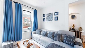 Una sala de estar con un sofá gris, cortinas azules y una ventana con vista a un edificio de ladrillo.