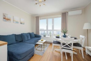 A modern living room with a blue sofa, a dining table with chairs, and a city view through large windows. - Spacious Apartment | Two Bedrooms and Air Conditioning (Warszawa)