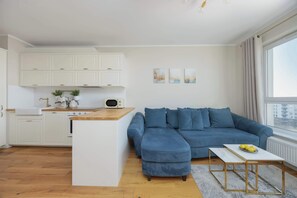 A modern living space with a kitchen, a blue sectional sofa, a small white table, and a window with curtains. - Spacious Apartment | Two Bedrooms and Air Conditioning (Warszawa)
