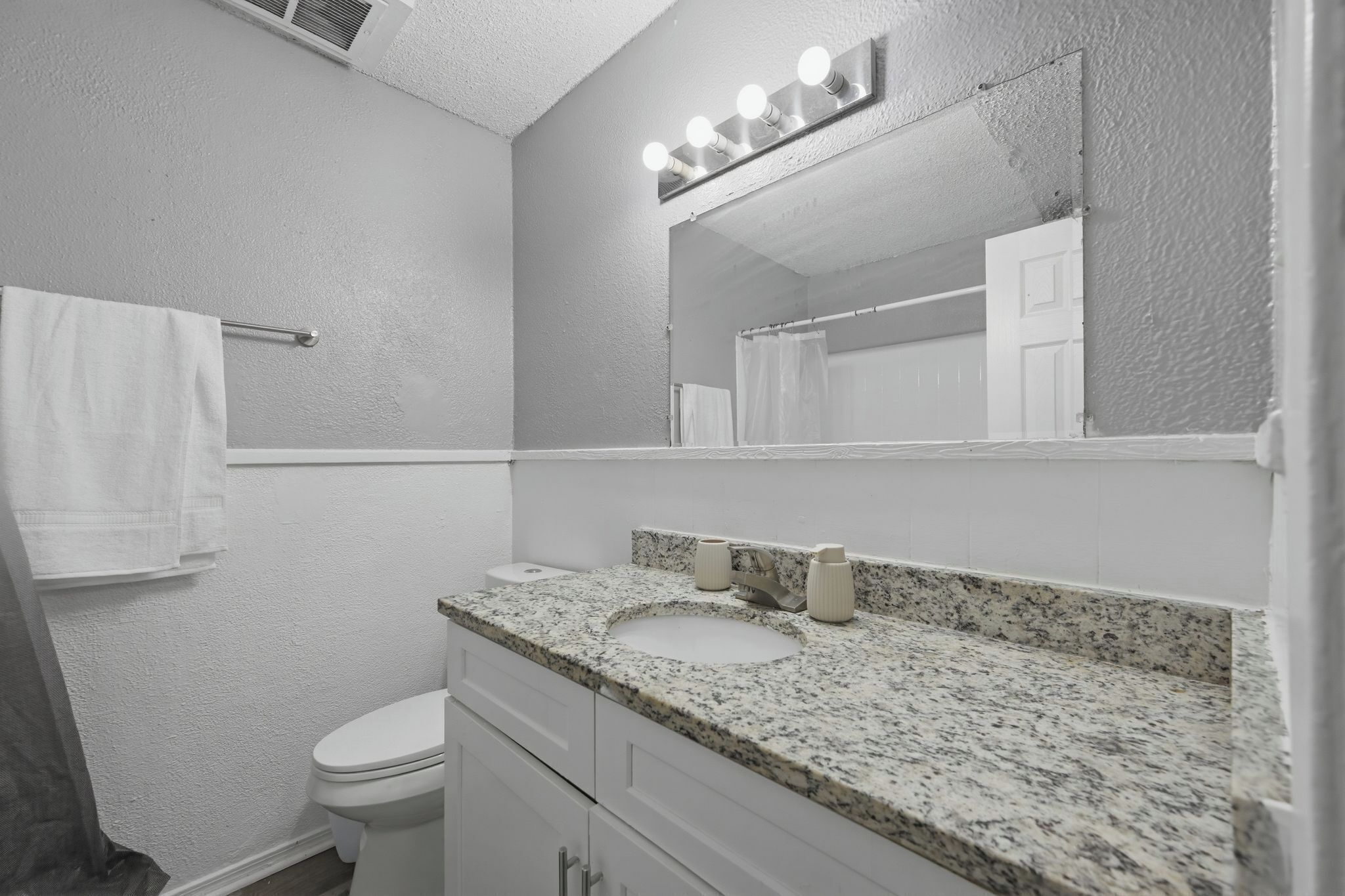 A bathroom with a white toilet, a granite countertop sink, a mirror with lights, and a towel rack.